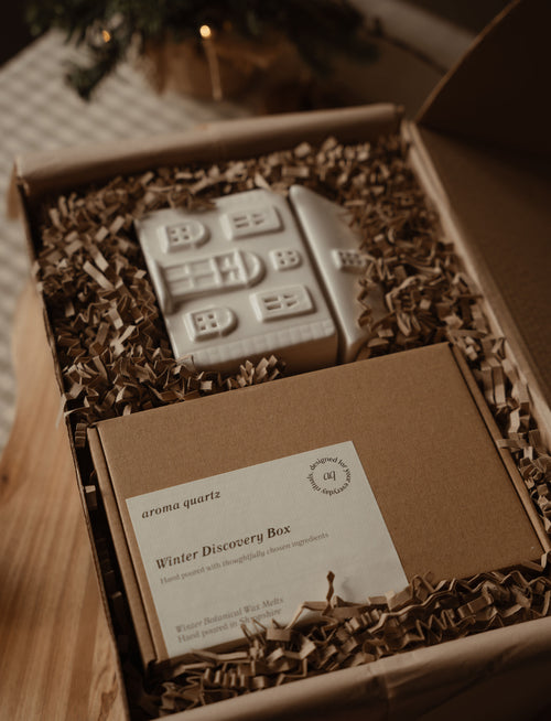 Open gift box with a ceramic house wax warmer and a Winter Discovery Box in shredded paper