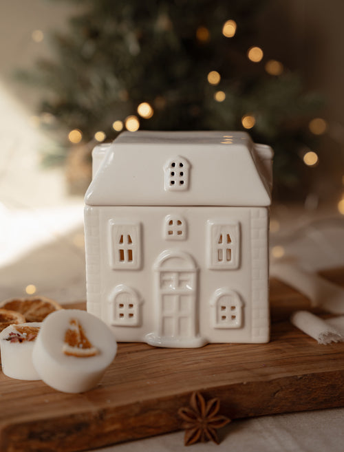 Ceramic house wax melt warmer with its roof on, beside winter wax melts on a wooden board