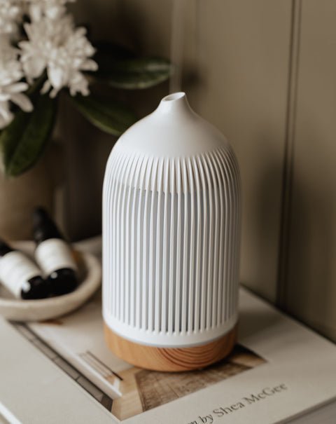 White diffuser with wooden base on a surface with flowers and bottles in the background