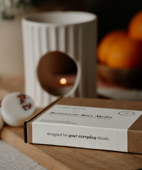 Box of botanical Wax Melts on a wooden surface with a candle and oranges in the background.