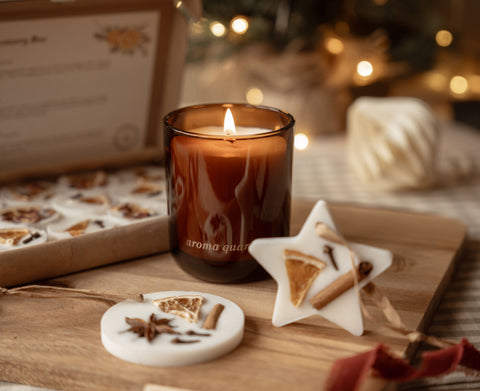 Lit amber candle with star and circle scented wax tree decorations beside the open box of winter wax melts