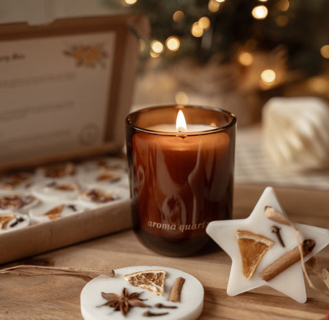 Close up of a lit amber candle and a star-shaped scented wax decoration with an open box of winter wax melts behind