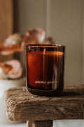Brown glass candle labeled 'aroma quinta' on a wooden surface with blurred seashells in the background.