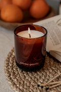 Lit candle in a brown glass container labeled 'aroma quartz' on a woven mat with oranges and an open book in the background.