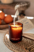 Candle labeled 'aroma quartz' with smoke rising, placed on a woven mat with pumpkins and an open book in the background.
