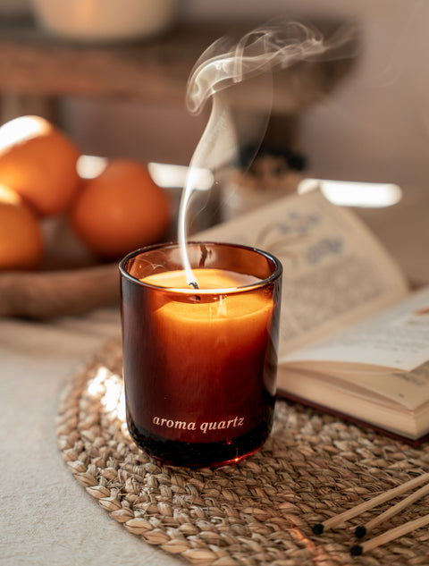 Candle labeled 'aroma quartz' with smoke rising, placed on a woven mat with pumpkins and an open book in the background.