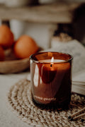 Candle in an amber glass with 'aroma quartz' text, surrounded by fruits and a book on a textured surface.