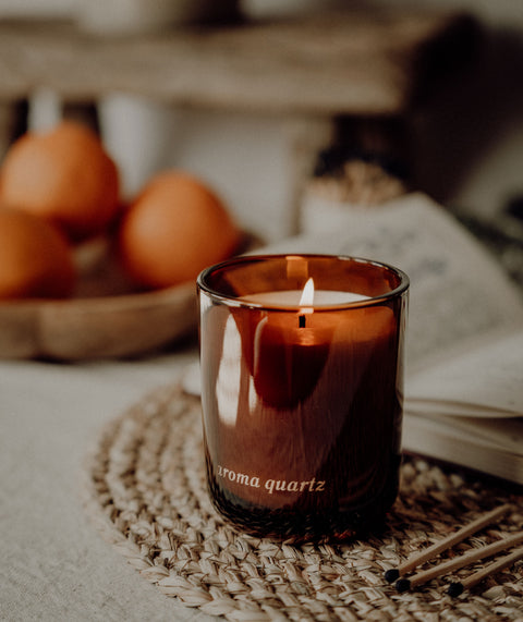Candle in an amber glass with 'aroma quartz' text, surrounded by fruits and a book on a textured surface.