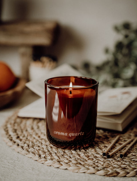 Candle in a glass jar labeled 'aroma quartz' on a woven mat with a blurred background