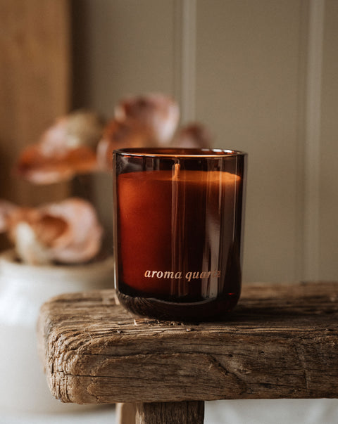 Brown glass candle labeled 'Aroma Quartz' on a wooden surface with blurred seashells in the background.