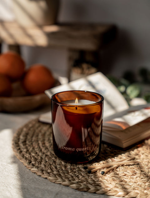Brown glass candle with 'Aroma Quartz' on a woven mat with books and oranges in the background