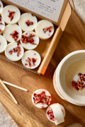 A flat lay style image of natural wax melts, each melt is finished with rose petals on a wooden surface.