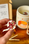 Hand holding a wax melt with dried flowers, with a blurred background of a wooden surface and a oil burner.