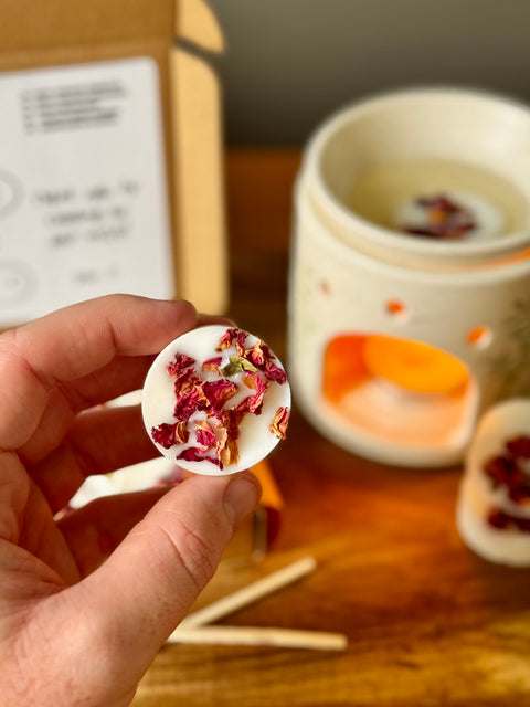 Hand holding a wax melt with dried flowers, with a blurred background of a wooden surface and a oil burner.