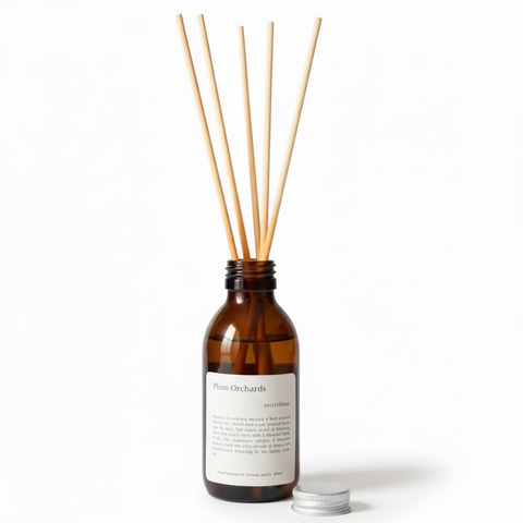 Brown diffuser bottle with wooden reed on a wooden surface and grey background, the cream coloured textured labels reads Plum Orchards Reed Diffuser.