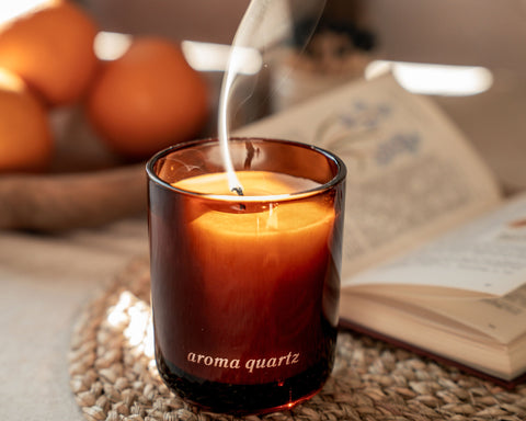 Candle labeled 'aroma quartz' with smoke rising, placed on a woven mat with oranges and an open book in the background.