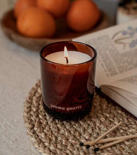 Lit candle in a brown glass container labeled 'aroma quartz' on a woven mat with oranges and an open book in the background.